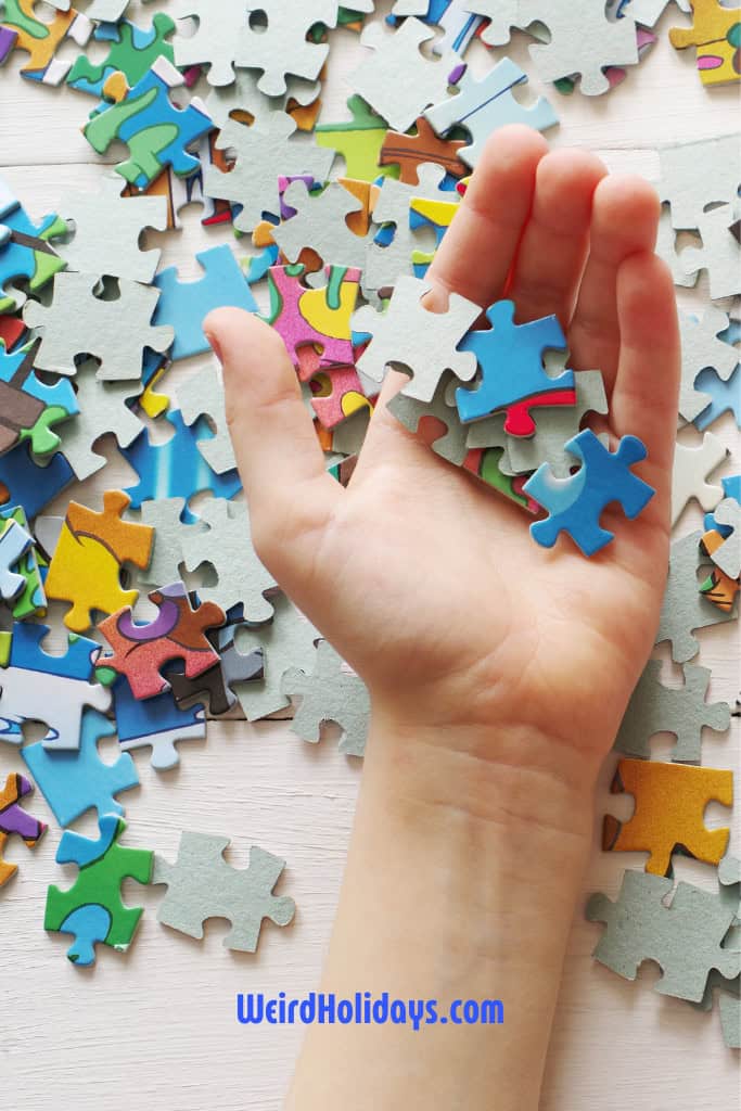 Alt text: A close-up of a hand holding several colorful jigsaw puzzle pieces above a scattered pile of puzzle pieces on a white wooden surface. The pieces are a mix of blue, green, red, and other vibrant colors. The text "WeirdHolidays.com" is displayed in blue at the bottom of the image.







